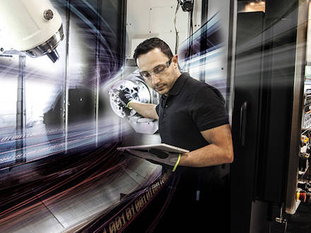 A man standing next to a CNC machine looking on a tabket that he holds in one of his hands